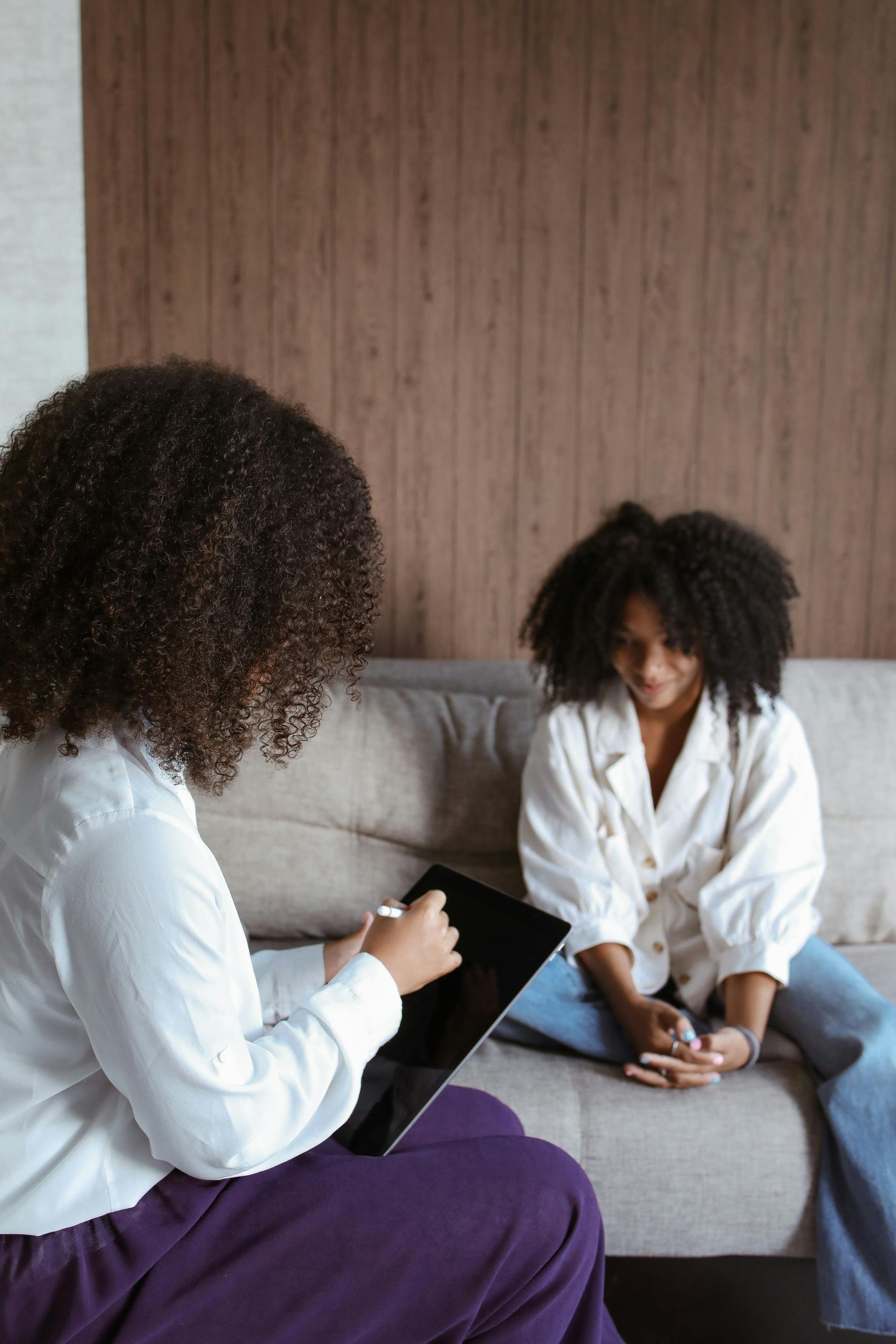 Two people in white coats talking on a couch, one holding a clipboard in a living room setting.