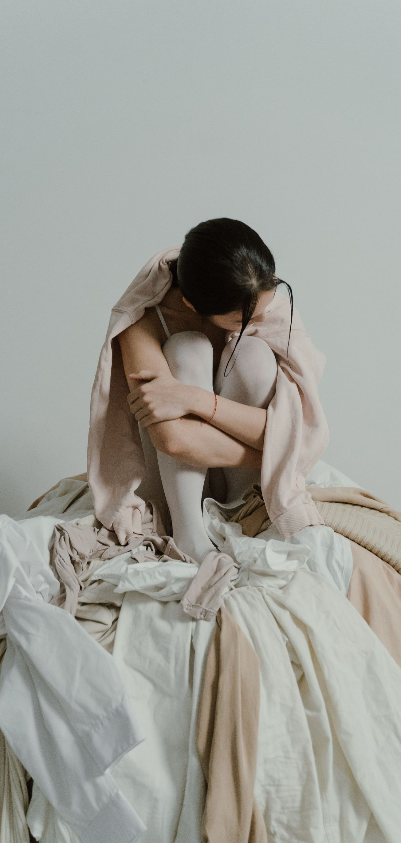 Person in a cream dress sits curled up on a bed against a plain wall.