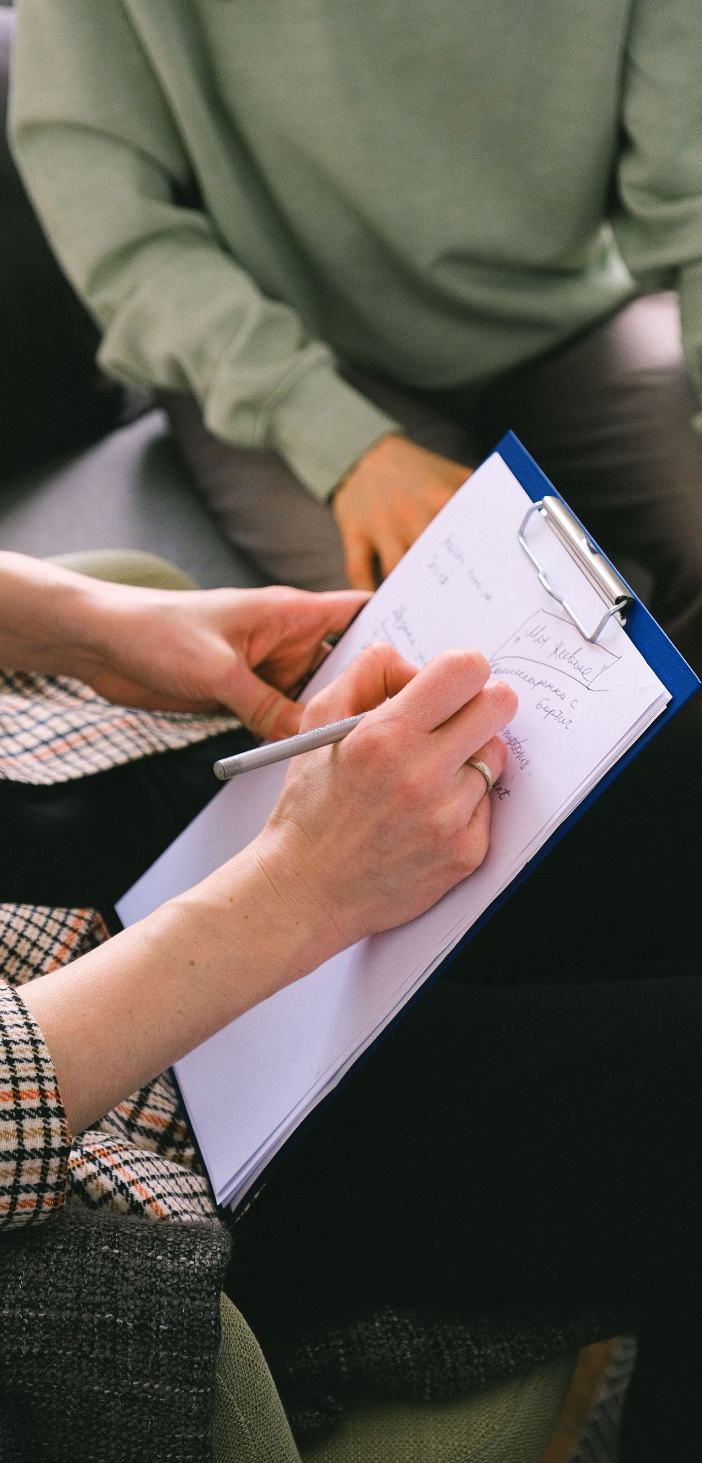 Person writing on a clipboard during a conversation, with another seated person in the background.