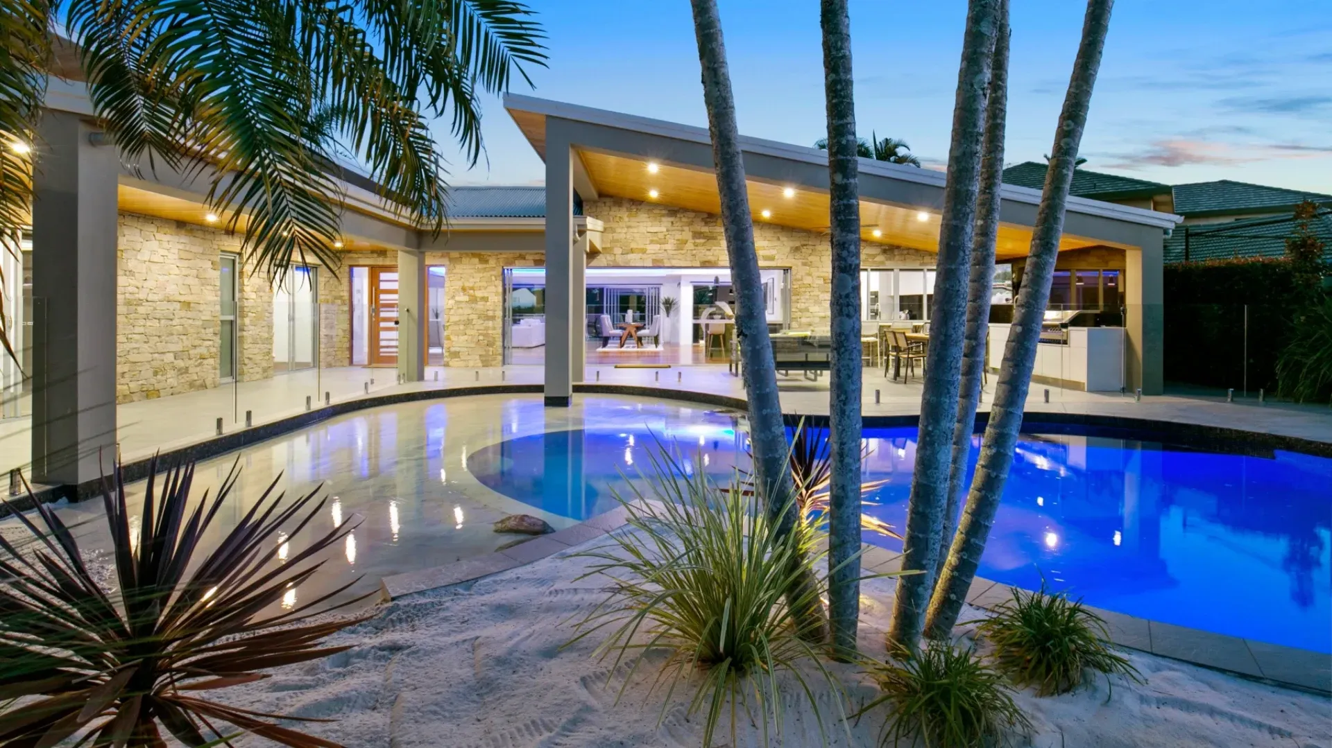 A modern house with a pool at dusk, palm trees, and sand. The pool is blue and the house has a slanted roof.