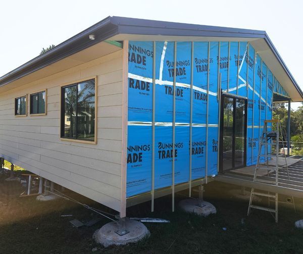House under construction; exterior partially covered with blue wrap, wood siding, and windows.