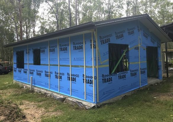 Building under construction, sheathed in blue wrap, with window and door openings.