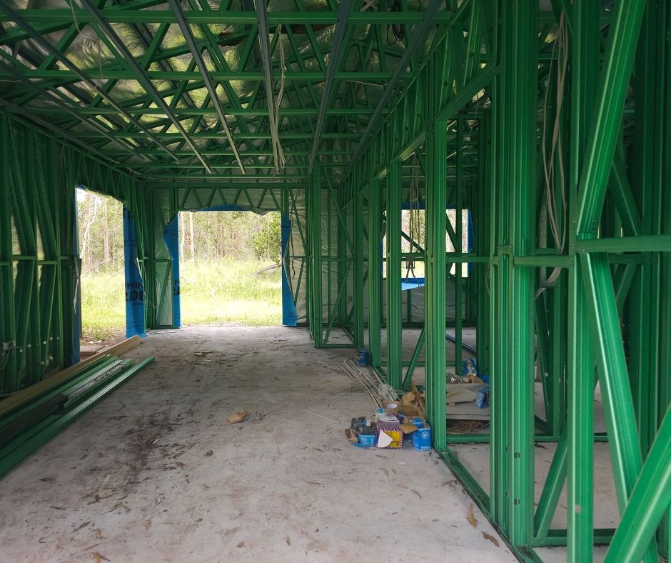 Interior view of a building frame under construction; green metal studs and ceiling beams, concrete floor, open doorway to outside.