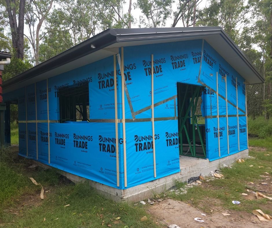 Small building under construction, wrapped in blue protective sheeting, with a forest background.