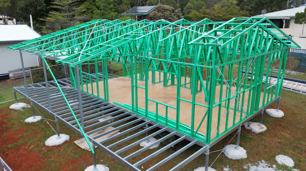 Green steel frame of a house under construction on a raised platform, set outdoors on grass.
