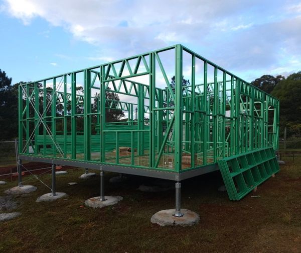 Green timber house frame under construction, elevated on steel posts. Set on a grassy area, cloudy sky in background.