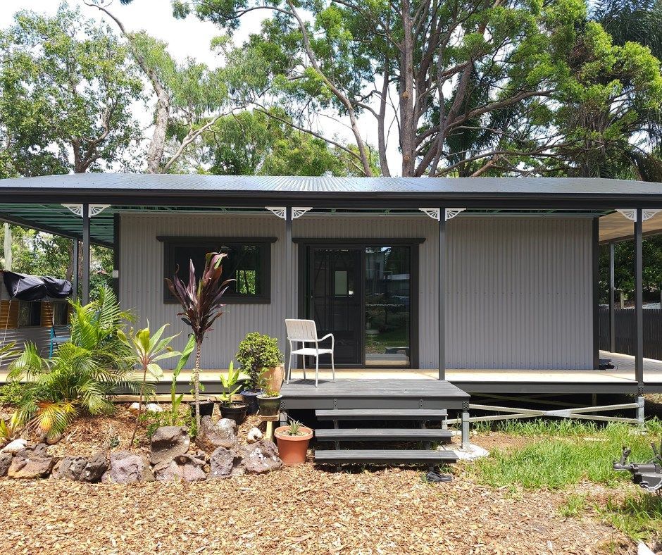 Gray house with covered porch, steps, and door; chair sits outside.  Plants and trees surround.