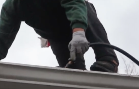 Person on roof, wearing gloves, working with a black hose on a gray gutter, overcast sky.
