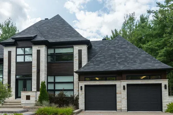 Modern two-story house with gray stone facade, dark roof, and two-car garage under a cloudy sky