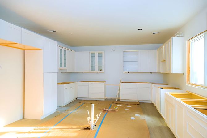 Bright unfinished kitchen with white cabinets, bare floors, and sunlight from a large window.