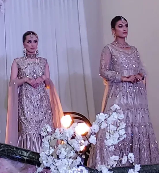 Two women in shimmering lavender gowns stand on a runway beside white floral arrangements.