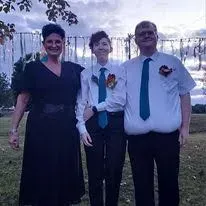 Three people standing outdoors at dusk, dressed in formal attire with boutonnieres.