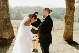Bride and groom exchange vows outdoors beneath trees in a sunlit ceremony