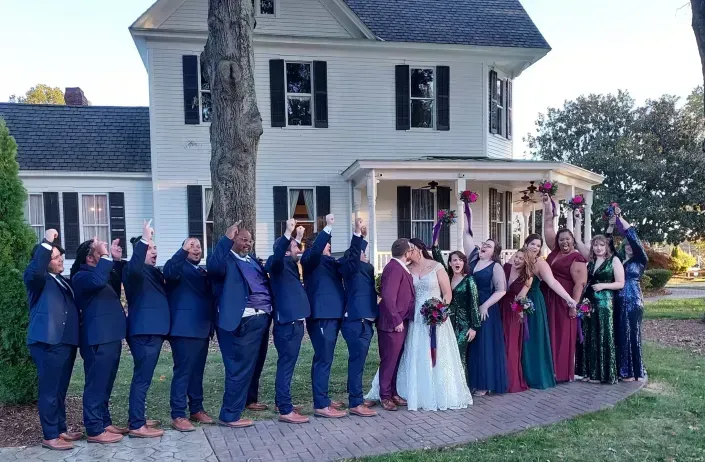 Wedding party posing in front of a white house, with bridesmaids in blue and colorful dresses and a bride in white.