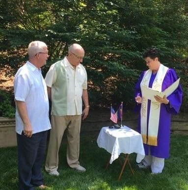 Outdoor blessing ceremony beside a small flag-draped table, with three people standing in a garden.