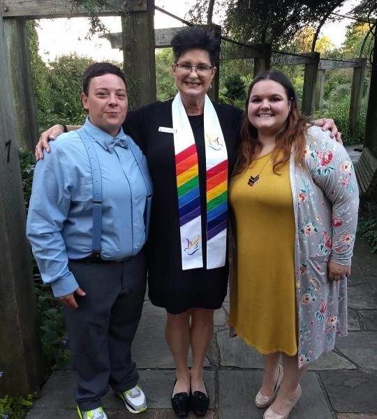 Three people pose outdoors; center person wears a rainbow stole and the others wear formal attire.