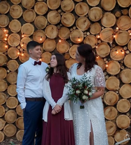 Three people posing in formal attire before a wall of stacked log ends with warm lights.