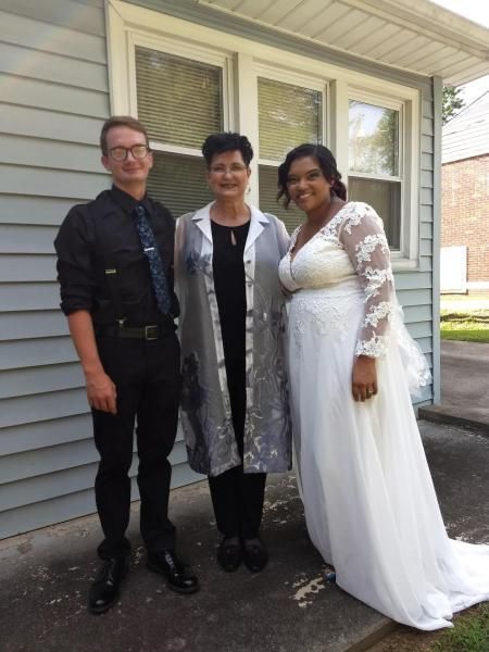 Three people standing together outside a house, smiling, with one in a white dress.