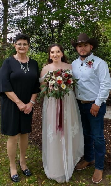 Bride in white dress holding bouquet stands with two guests outdoors in front of trees.