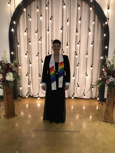 Person standing indoors before a white draped arch with hanging lights and floral arrangements.