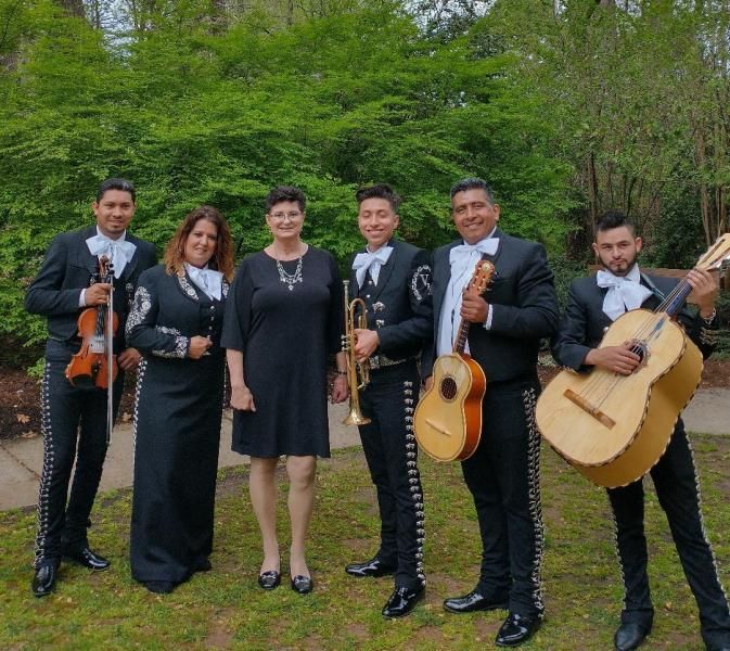 Six musicians in black formalwear pose outdoors with violins and guitars in a green park.
