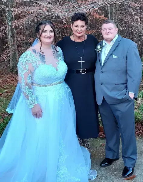 Three people posing outdoors at a wedding, with the bride in a light blue gown and the officiant in black.
