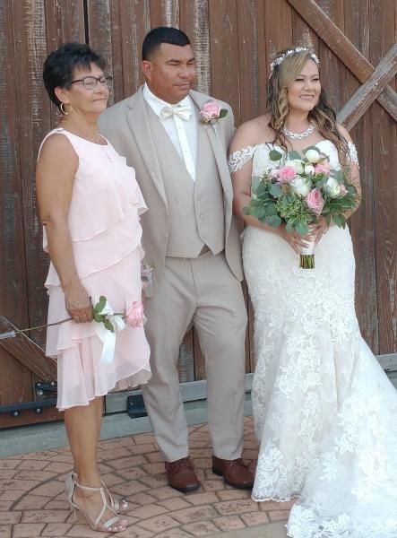 Three people posing at a wedding, with the bride holding a pink-and-white bouquet in front of wooden doors.
