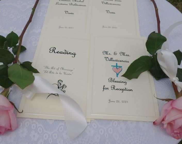 Wedding program on a cream fabric table, decorated with white and pink roses and white ribbon.