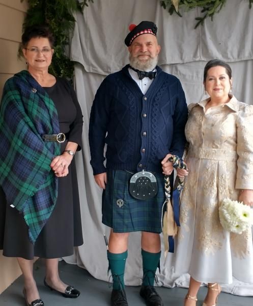 Three people in formal Scottish attire posing indoors, with floral decorations behind them.
