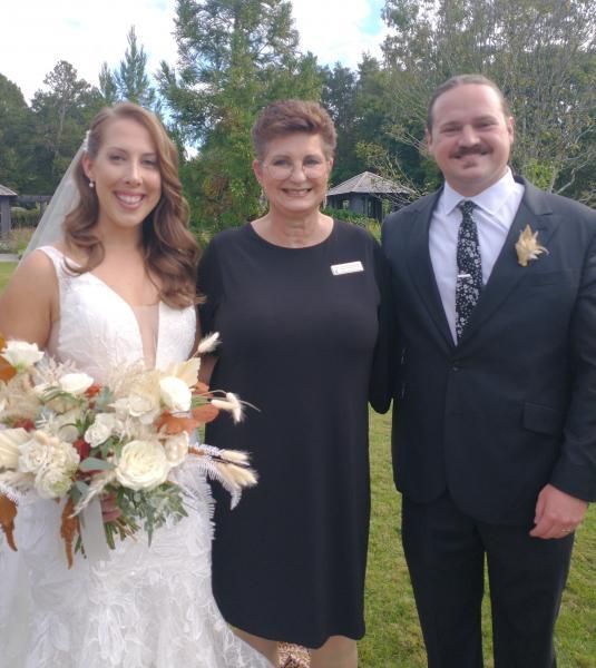 Three people posing outdoors, including a bride holding a white bouquet and two formally dressed guests.