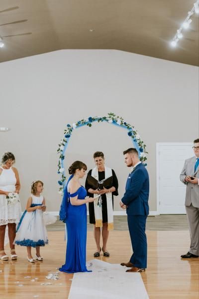 Small indoor wedding ceremony with couple exchanging rings under floral arch, guests nearby