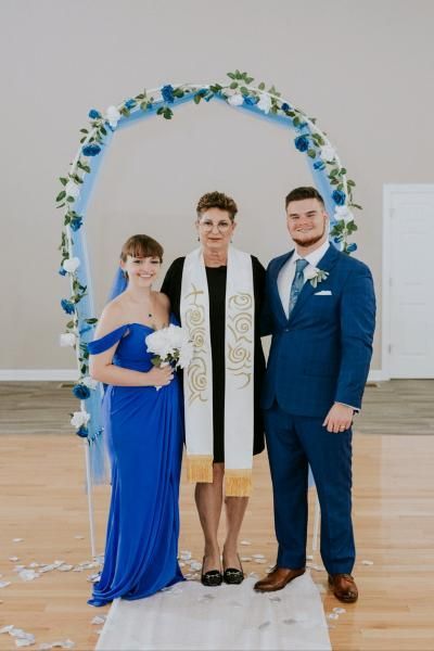 Three people pose under a blue-and-white floral arch at a wedding venue