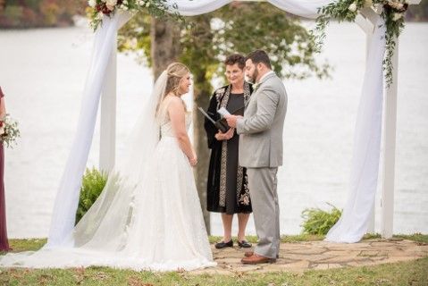 Bride and groom stand with an officiant beneath a floral arch by the water.