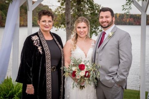 Three people posing outdoors by the water, with a bride holding a bouquet in the center.