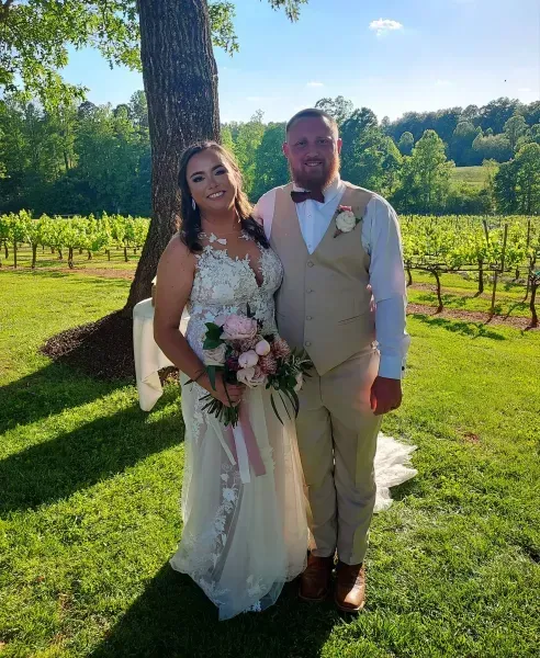 Wedding couple standing outdoors in sunlight, smiling and holding a bouquet in a vineyard.