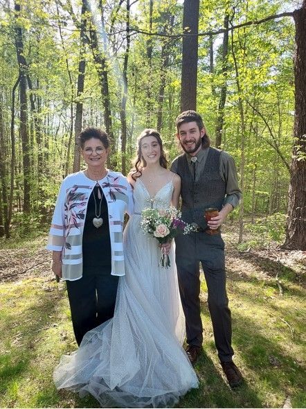 Three people pose in a sunlit forest, including a bride in a white dress holding a bouquet.