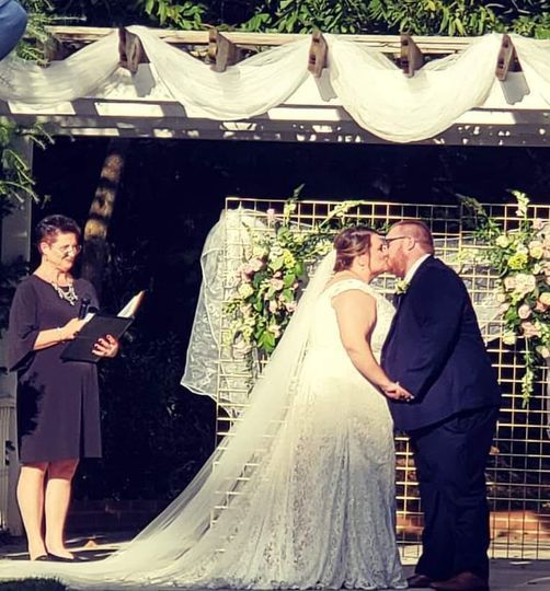 Wedding ceremony under a pergola, bride and groom kiss as officiant stands nearby.