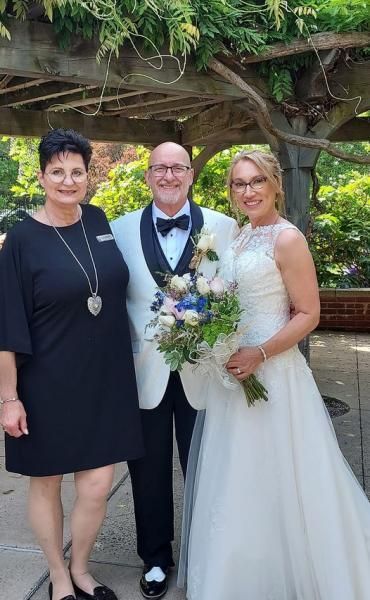 Three people pose outdoors at a wedding, with the bride in a white dress and the groom in a tuxedo.