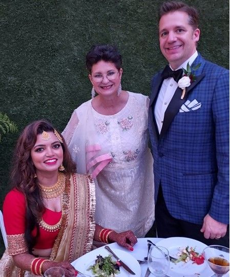 Three people dressed formally pose at a wedding table with flowers and drinks.