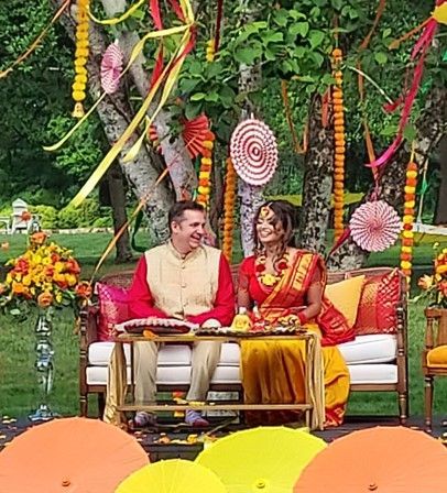 Couple seated on a decorated outdoor stage, smiling in traditional attire amid colorful hanging decorations.