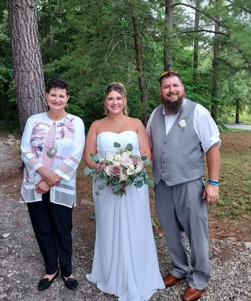 Bride and two guests stand outdoors in a wooded area, posing for a wedding photo.