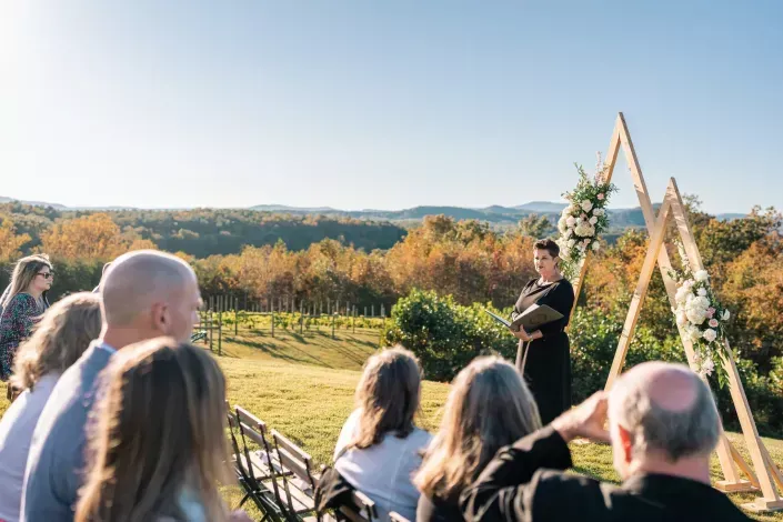 Outdoor wedding ceremony at sunset, with guests seated near a floral arch and speaker facing them