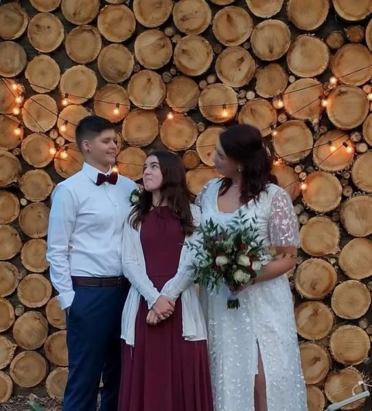 Three people in formal attire posing in front of a wall of stacked logs with string lights