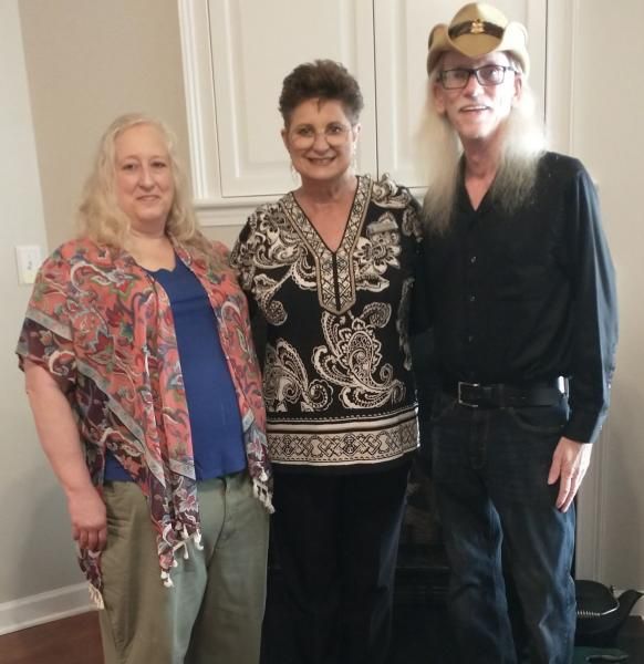 Three people standing indoors, smiling at the camera; two wear patterned tops and one wears a black outfit and hat.