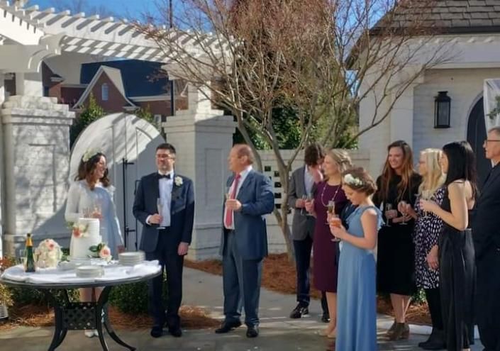People mingling at an outdoor gathering by a white house and pergola, holding drinks and chatting.