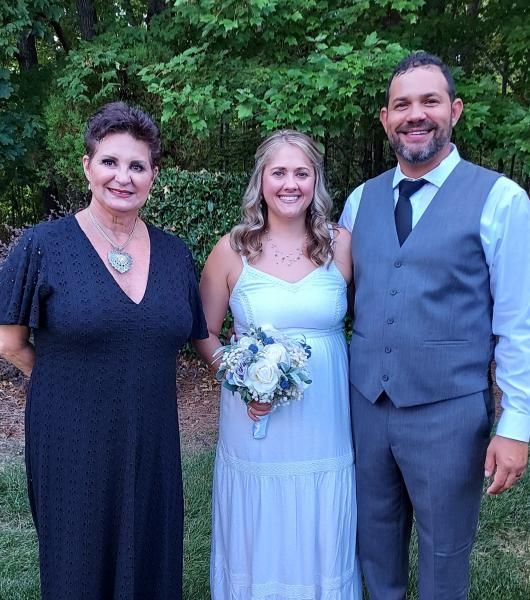 Three people posing outdoors, with a bride holding a bouquet between two formally dressed companions.