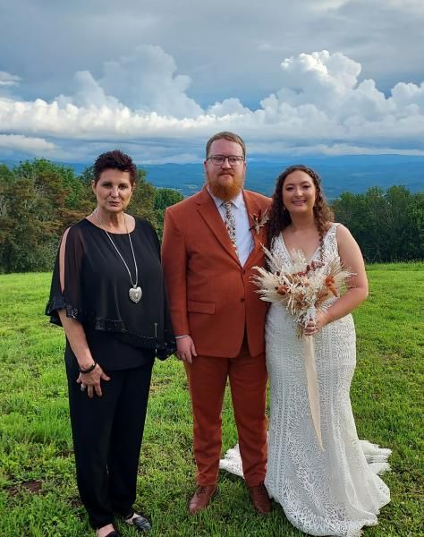 Three people standing outdoors at a wedding, with mountains and clouds in the background.