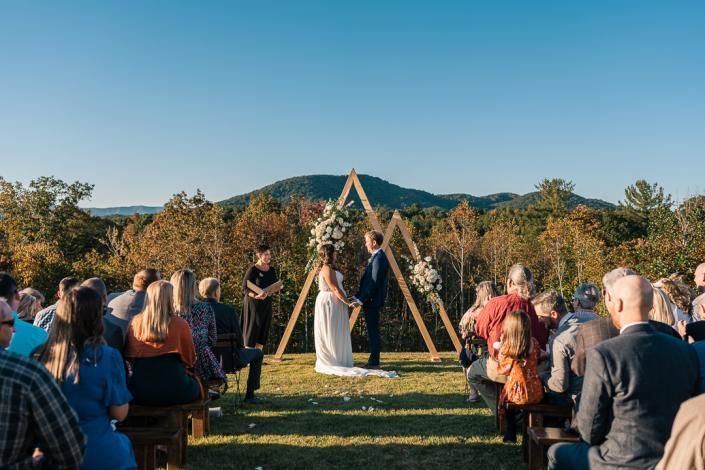 Outdoor wedding ceremony under a wooden arch with guests seated on both sides, mountains in the background