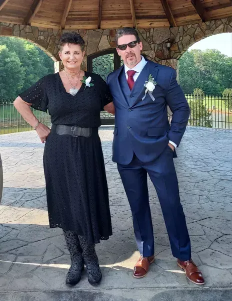 Two people dressed formally standing together under a wooden gazebo, smiling outdoors.