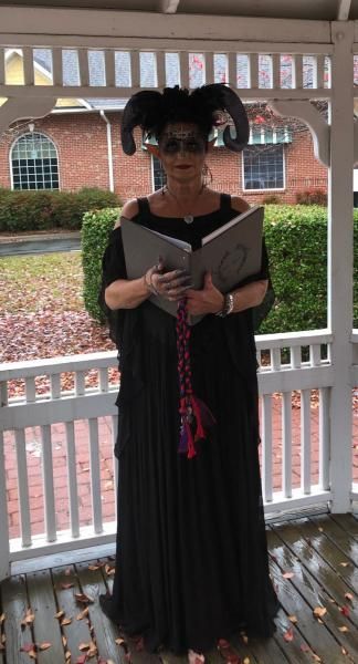 Person in a black dress and horns holds an open book on a porch with brick building in the background